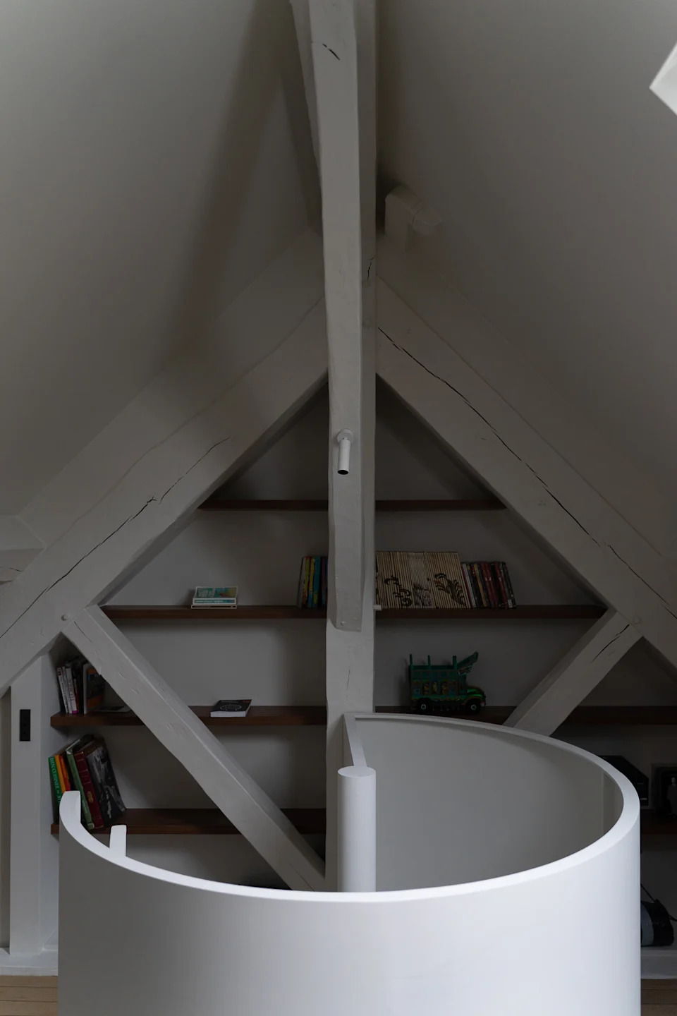 A refined attic apartment interior characterized by a warm, modernist aesthetic. The space is organized as an open-plan living area under a pitched roof, with exposed white-painted beams and triangular dormer windows that bring in soft natural light.