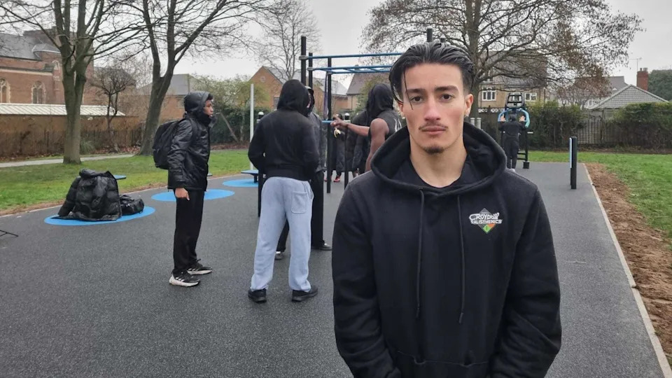 Theo Dopson wearing a dark hoodie stands in the foreground of an outdoor fitness area. Behind this person, several others are gathered around metal workout bars. The ground is covered with rubber safety surfacing, and leafless trees and nearby houses can be seen in the background on an overcast day.
