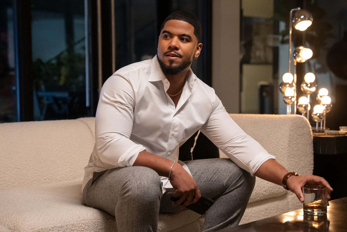 A man in a white shirt sits on a modern couch in a stylish, warmly-lit living room, holding a glass. The setting feels relaxed and sophisticated, with ambient lighting and contemporary decor.