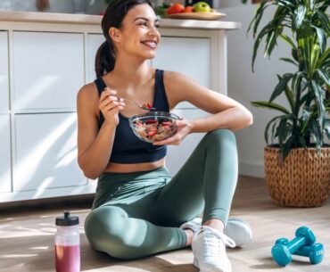 Shot of athletic woman eating a healthy bowl of muesli with fruit sitting on floor in the kitchen at home