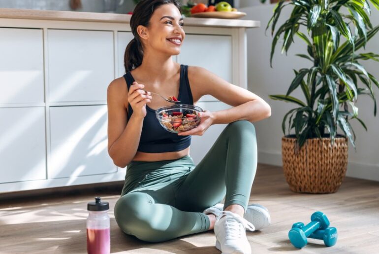 Shot of athletic woman eating a healthy bowl of muesli with fruit sitting on floor in the kitchen at home