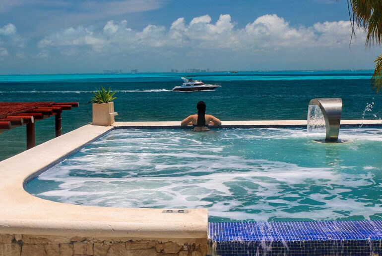 Luxury infinity pool with waterfall feature overlooking the crystal blue waters in a tropical paradise. Heather from Better Living enjoys the serene view of the ocean with a yacht sailing in the distance and palm trees framing the scene. This image embodies the travel and wellness lifestyle promoted by Better Living blog. Photo by Eric Weiss.
