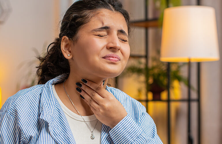 A woman holding her throat in pain while sitting at her desk at home.
