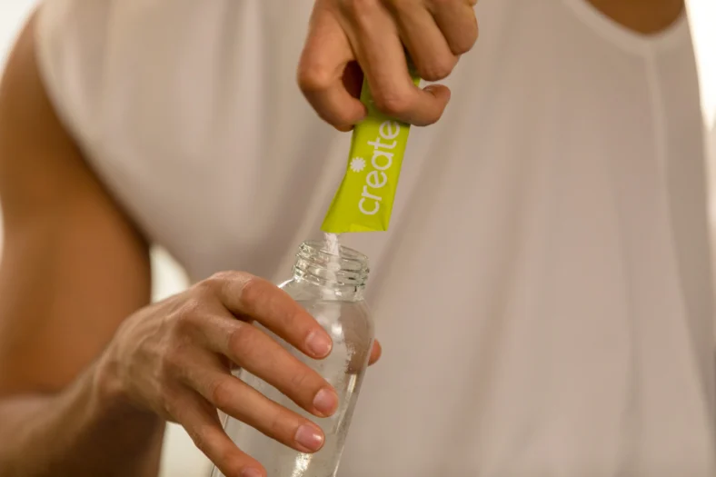 Person pouring Create electrolyte powder stick into a reusable water bottle for hydration and fitness recovery.