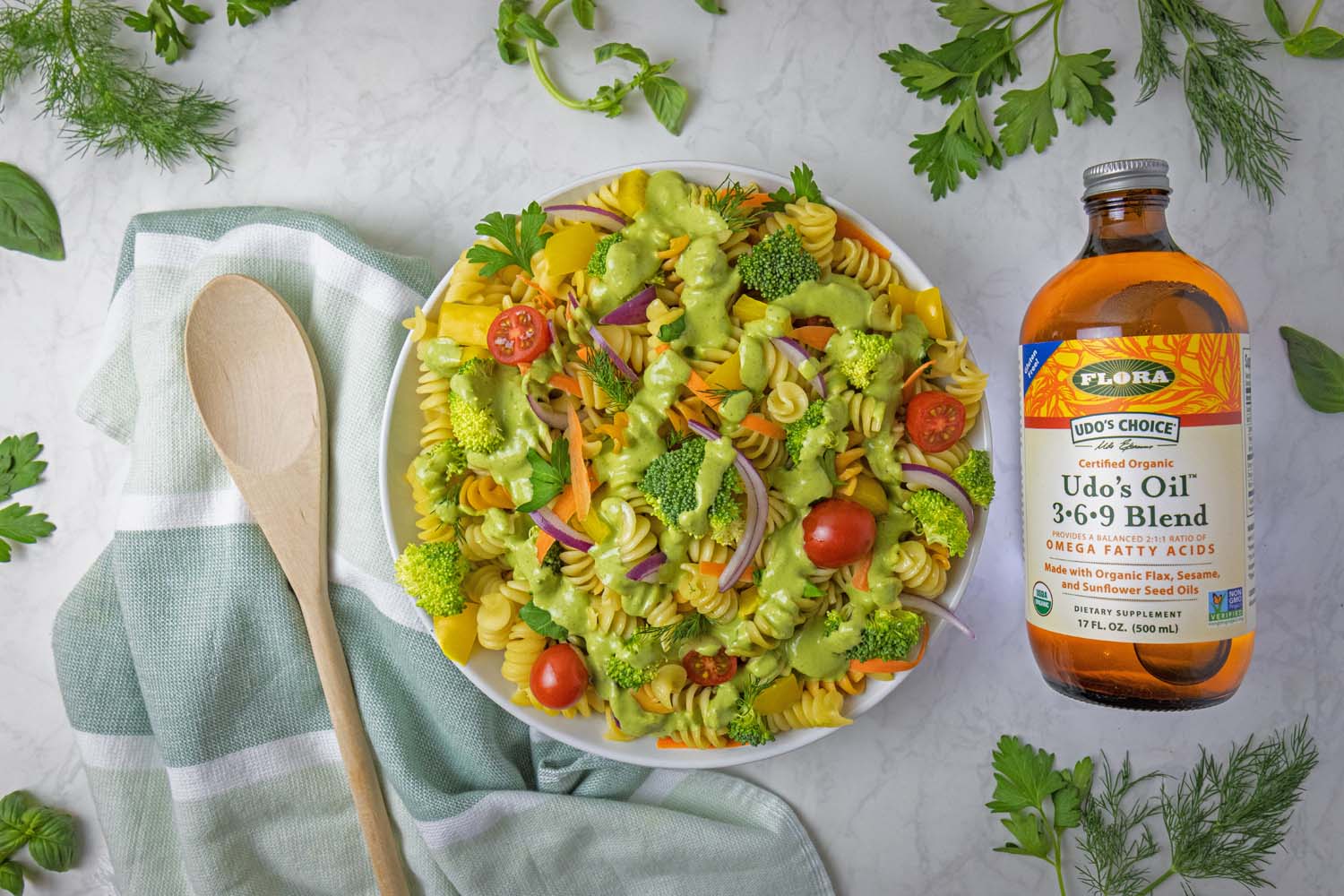 A beautifully styled overhead shot of completed Green Goddess pasta salad in a white bowl, surrounded by fresh herb sprigs including parsley, dill, and basil scattered around a white marble surface. A bottle of Udo's Oil 3-6-9 Blend sits to the right, with a wooden spoon resting on a sage green striped kitchen towel to the left. The pasta salad shows vibrant colors from the tri-color rotini, green broccoli, red cherry tomatoes, yellow bell peppers, and purple onions.