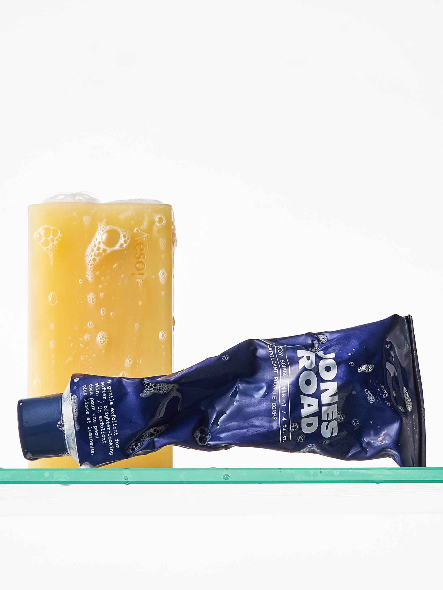 An Aesop body soap and Jones Road body scrub on a glass shower shelf