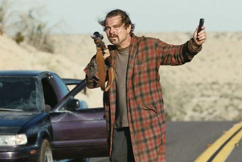 A man in a plaid robe stands on a deserted road holding a rifle and a handgun, with a car parked behind him and desert landscape in the background.
