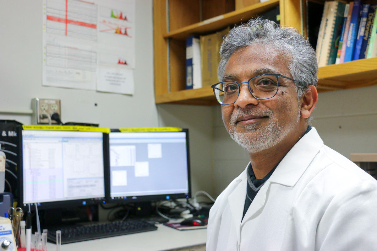 M. Suresh sits at a computer desk and smiles to someone off camera. He is wearing glasses and a white lab coat.