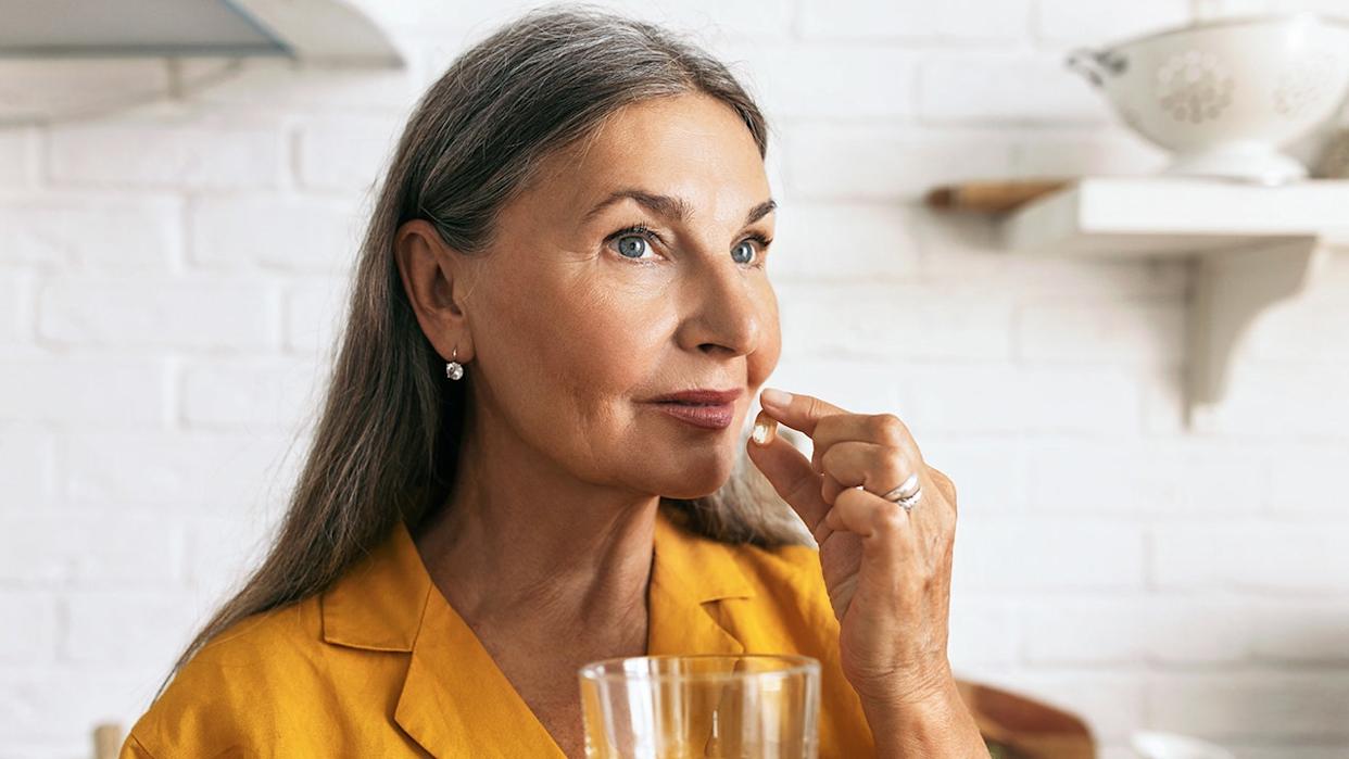 Older woman holding a capsule pill near her mouth with a glass of water in hand.