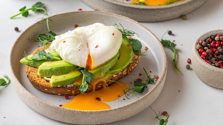 Avocado toast with poached egg and micro greens on gray ceramic plate next to bowl of peppercorns