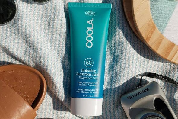 A tube of blue and white sunscreen is placed on a white cloth alongside a brown leather accessory, a wooden plate, and a camera.