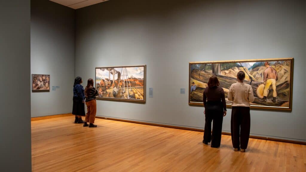 Four people view large framed paintings on the walls of an art gallery with wooden floors and neutral-colored walls.