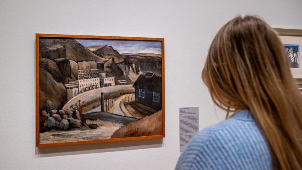 A person with long hair views a framed painting of an industrial landscape displayed on a gallery wall.