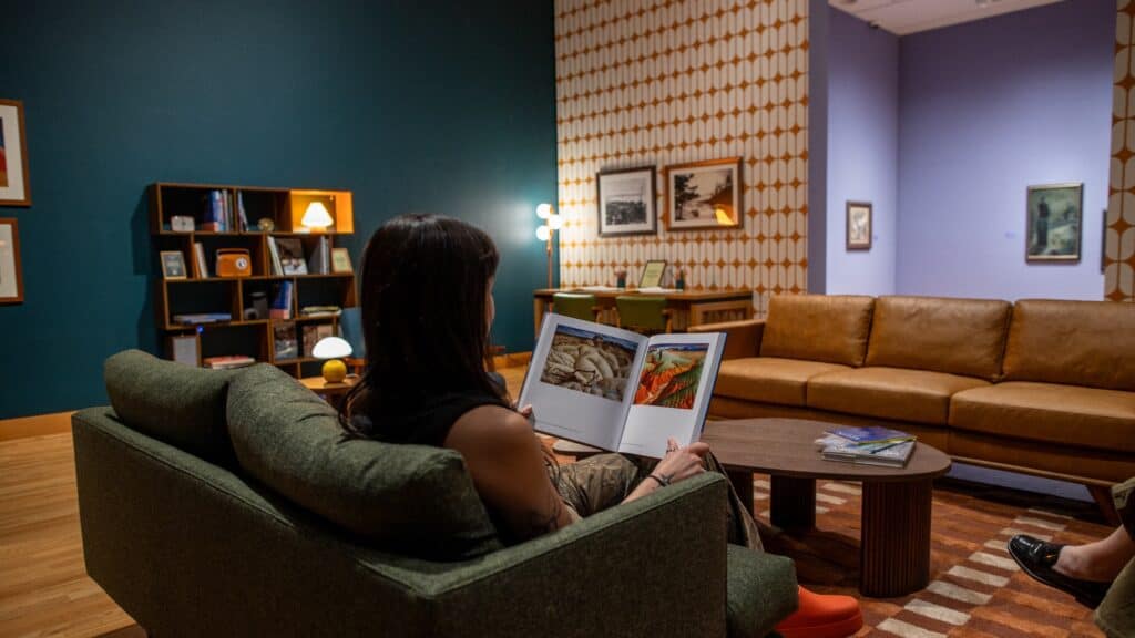 A person sits on a green sofa reading a book in a retro-style living room with patterned wallpaper, wooden furniture, and framed pictures on the walls.