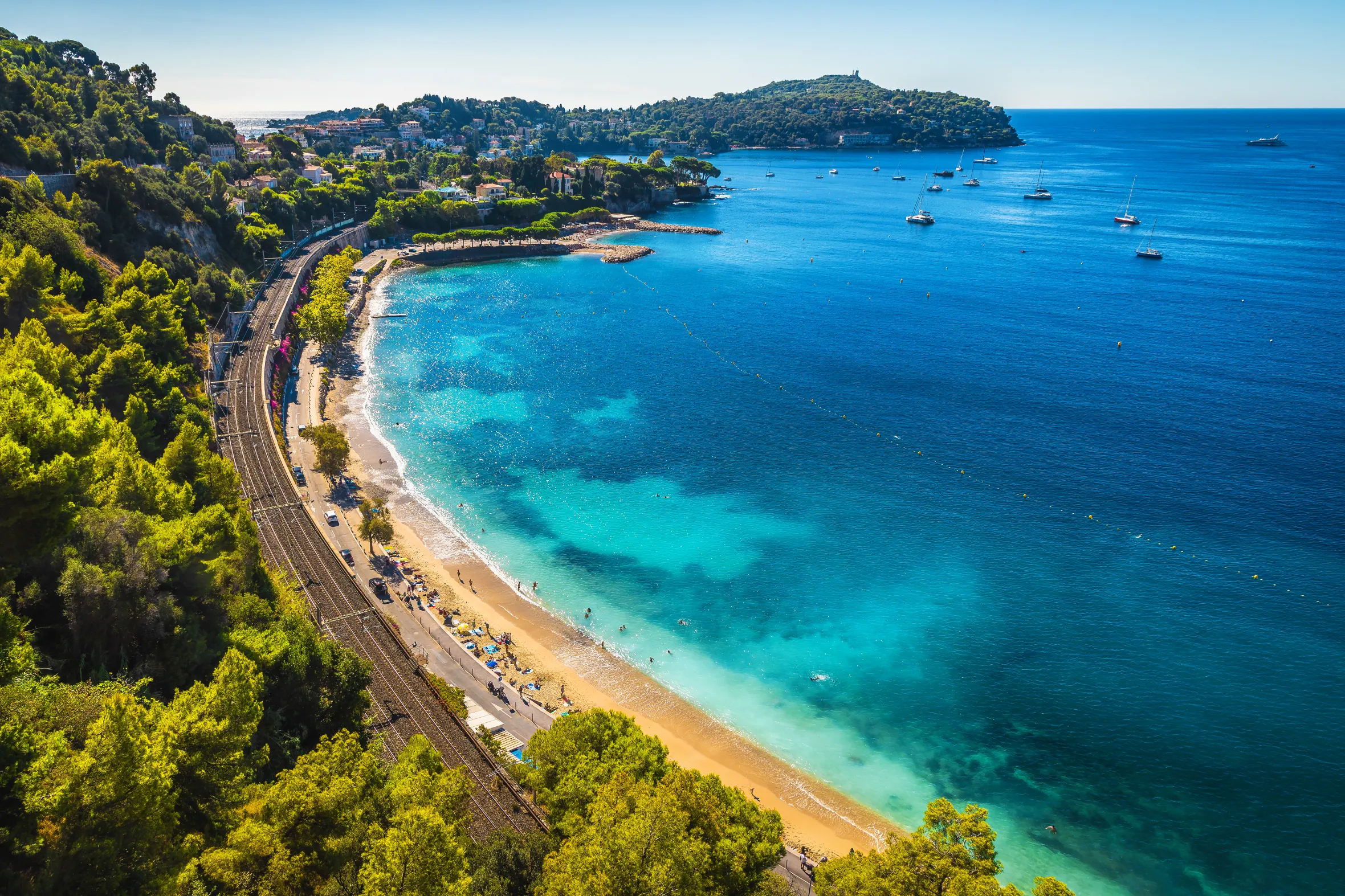 Aerial view of Villefranche sur Mer with its turquoise bay, sandy beaches, coastal town, and a train line running along the shore.