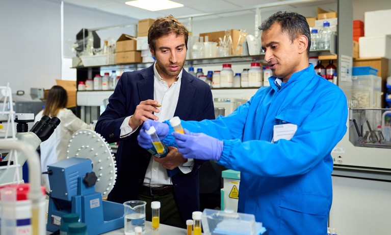 A laboratory setting with two individuals standing at a workbench surrounded by scientific equipment. One individual wearing a blue lab coat and gloves is holding up two sample containers for examination. The other individual, dressed in a dark blazer, is observing. The workspace includes machines, glass beakers, bottles, and shelves stocked with laboratory supplies in the background.