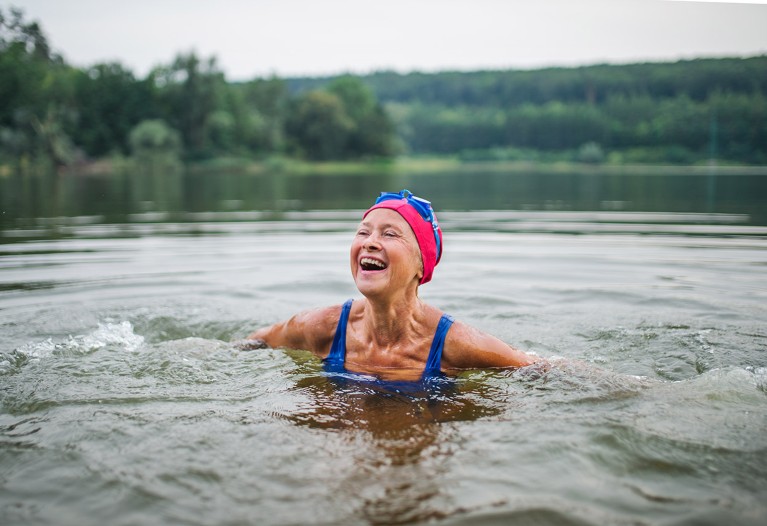A senior woman in a swimsuit and swimming cap laughs while enjoying a wild swim in a large outdoor lake.