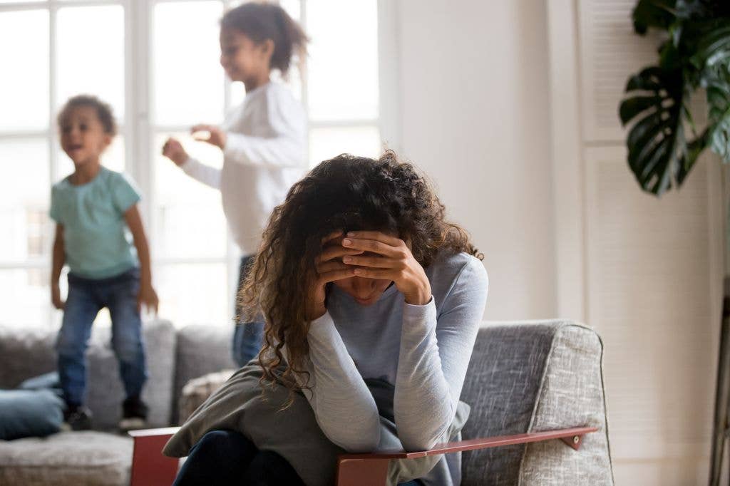 Black single frustrated woman hold her head with hands sitting on chair in living room, playful kids jumping on couch on a background. Tiredness, depression difficult to educate children alone concept