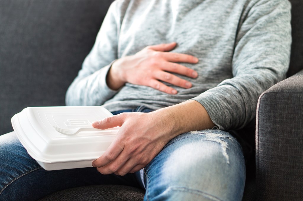 Man in jeans and a gray sweatshirt rubbing his stomach while holding a white takeout box with a plastic fork on his lap.