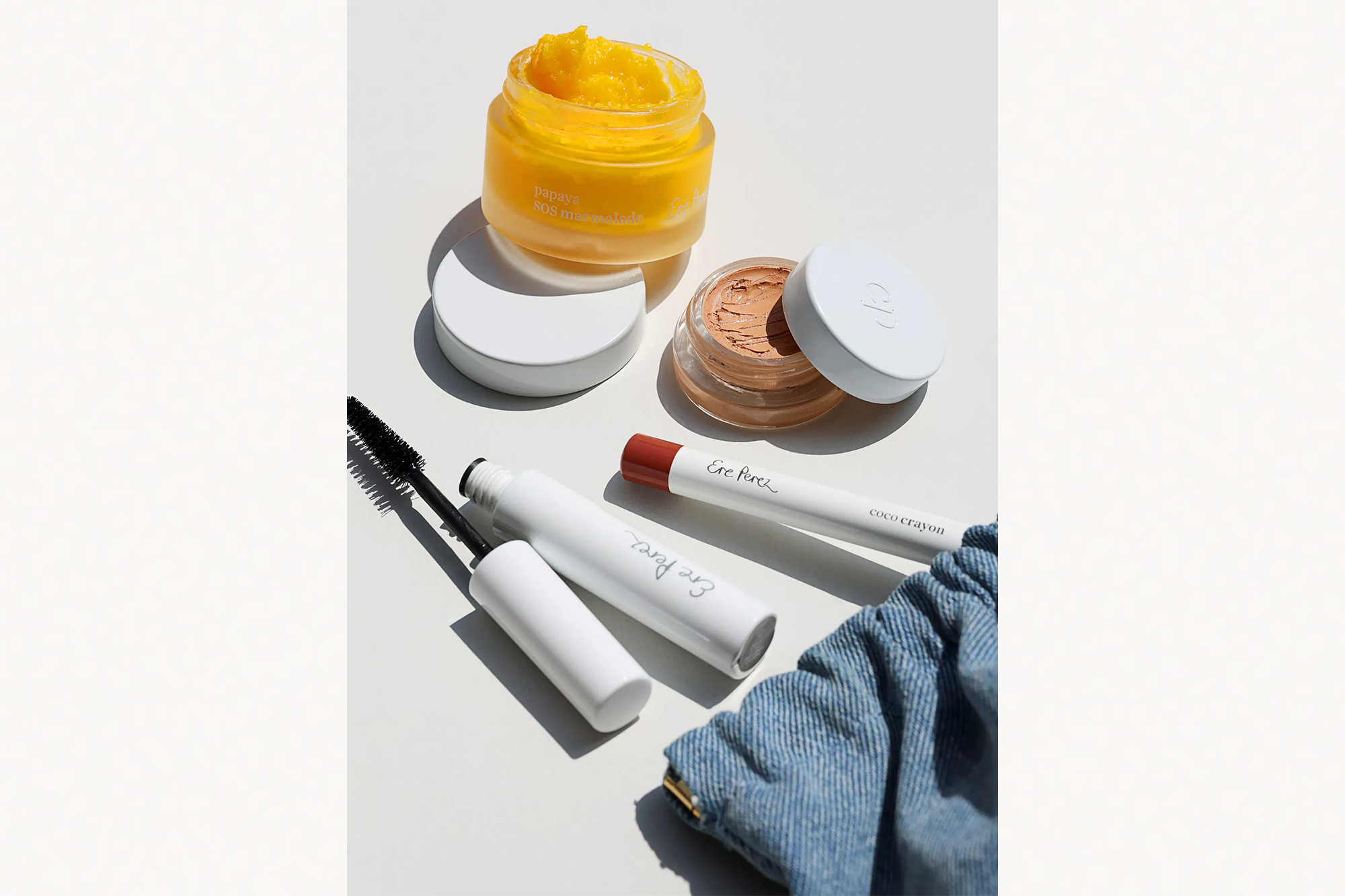 Ere Perez Papaya SOS Marmalade, Coco Crayon, and mascara arranged on a white surface next to a denim cosmetic bag.