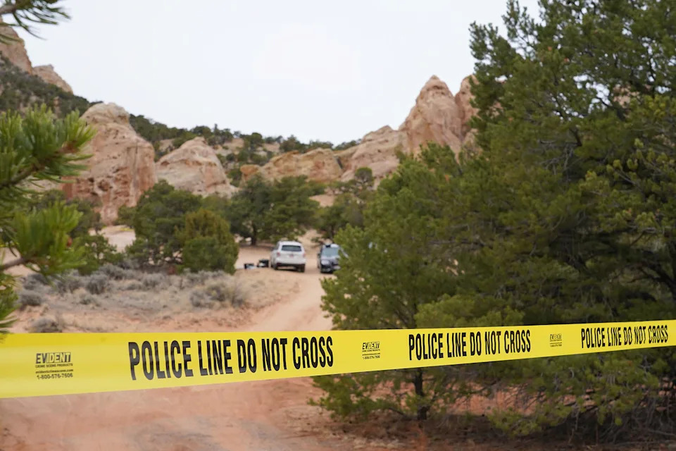 Yellow police tape stretches across a red dirt road leading toward two parked vehicles, surrounded by small jagged rock formations. (George Frey / AP)