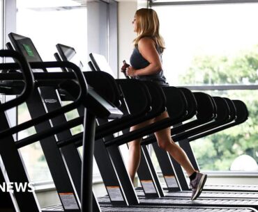 A woman wearing gym gear runs of a treadmill near large glass windows with trees outside. There are several empty treadmills next to her.