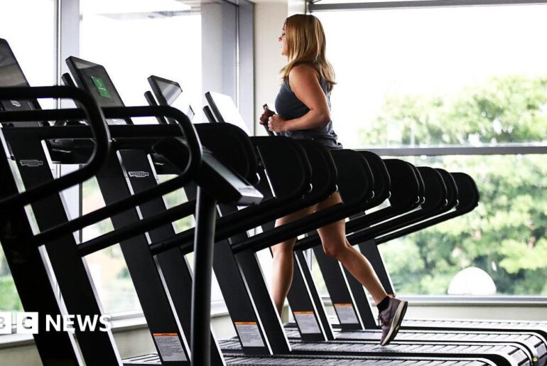 A woman wearing gym gear runs of a treadmill near large glass windows with trees outside. There are several empty treadmills next to her.