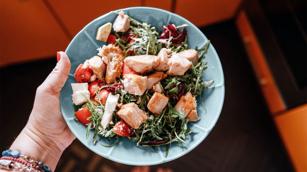 A person holds a bowl of salmon salad with greens and tomatoes, to represent healthy fats