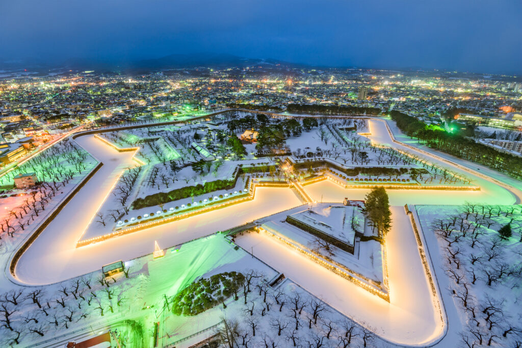 Aerial view of Goryokaku, a star-shaped fort in Hakodate, Japan, illuminated at night and surrounded by snow-covered grounds.