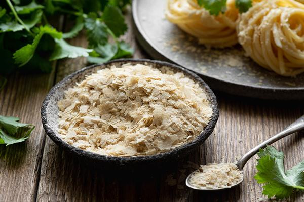 A ceramic bowl filled with nutritional yeast flakes on a wood surface, with a spoon containing some of the flakes next to the bowl.