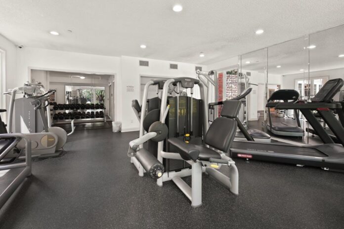 A well-lit indoor gym features treadmills, weight machines, and a rack of dumbbells in the background, all set on a black rubber floor.