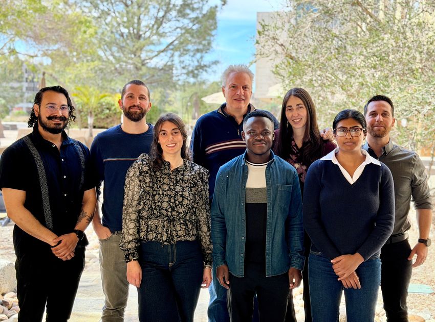 A photograph of adult men and women standing outside with a blurred background of trees for a photograph. Tatiana Hurtado de Mendoza (second from right in back), a cancer biologist at the University of San Diego, has long dark hair. Chris Benedict (back, left of Hurtado de Mendoza), a virologist at the La Jolla Institute for Immunology, has short, light-colored hair.