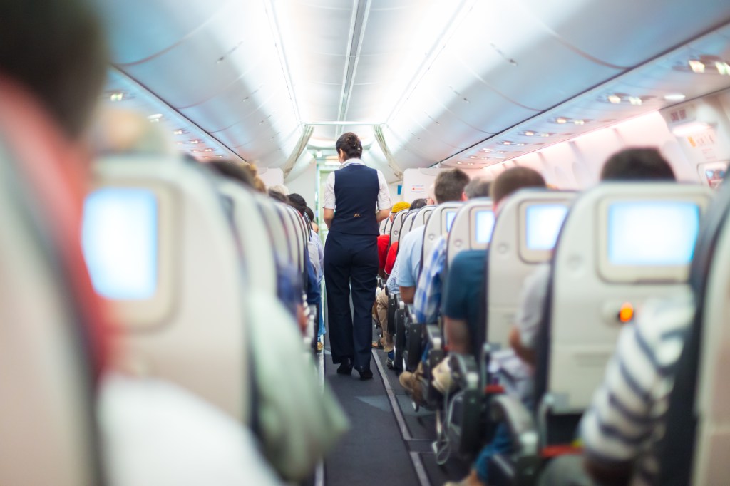 An airline stewardess in uniform walks down the aisle of an airplane filled with seated passengers.