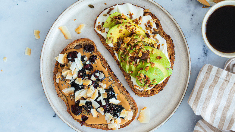 Toast with cream cheese, avocado, and seeds next to toast with peanut butter, blackberries, and coconut on white plate