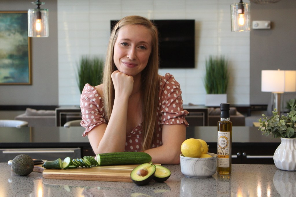 Kate Regan sits at a kitchen counter with a cutting board of sliced cucumber and avocado, with lemons and a bottle of olive oil nearby, smiling toward the camera.