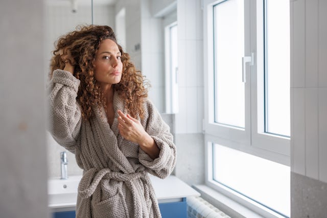 Mid adult woman with curly hair wearing a soft bathrobe stands thoughtfully in a bright bathroom, fi...
