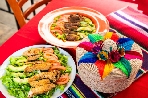 Grilled chicken and steak salads with avocado and tomatoes on a red table beside a colorful woven basket.