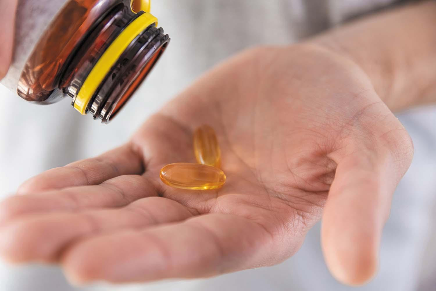 A hand holds two vitamin D supplement capsules just tipped out of a bottle held above the hand.