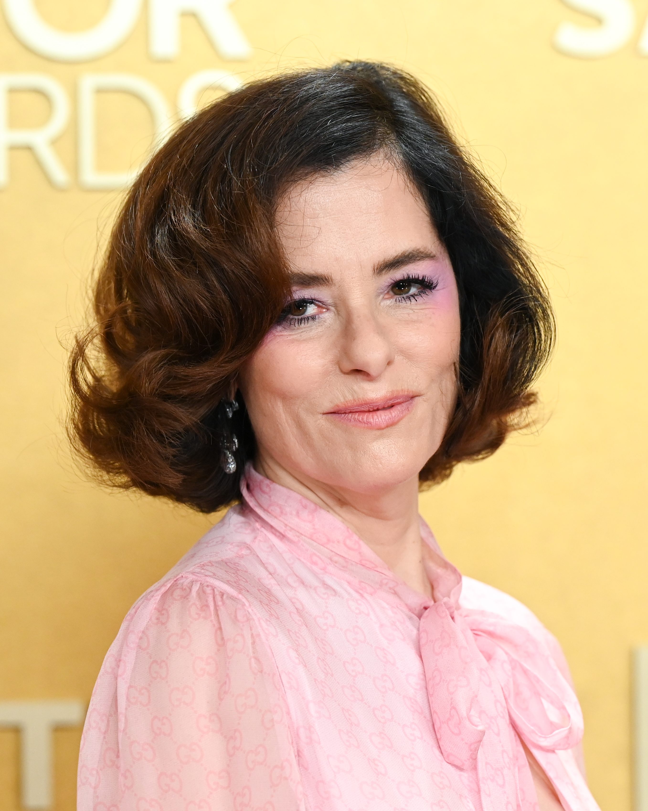 a white woman wearing a pink dress and pink eye makeup posing in front of a yellow backdrop on a red carpet