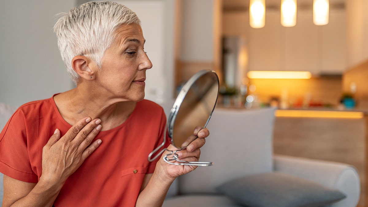 Short haired elderly woman holding mirror and applying face cream at home.