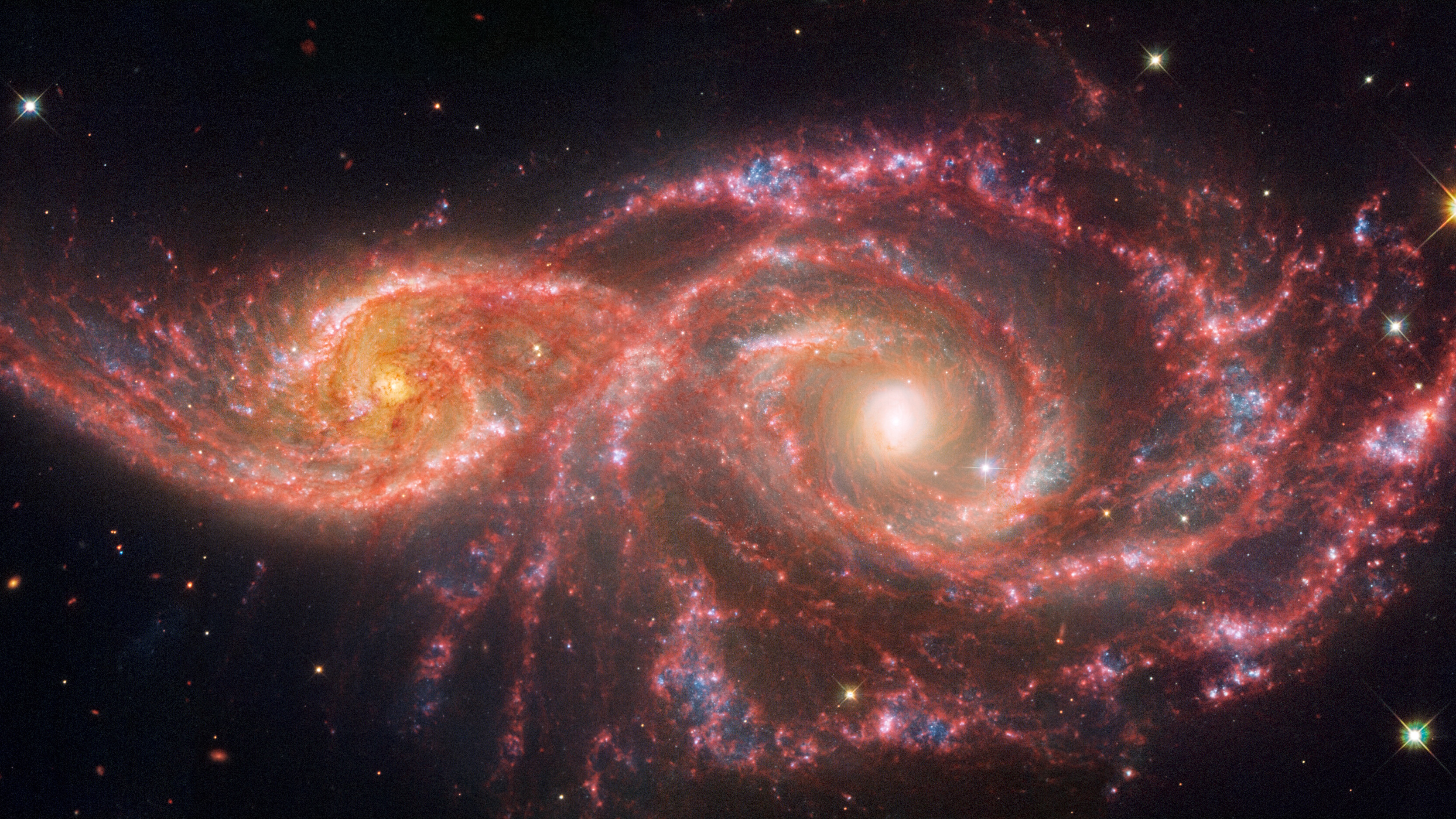 Two colorful spiral galaxies interacting in space, with bright centers and swirling arms of red, blue, and white hues, set against a backdrop of stars.
