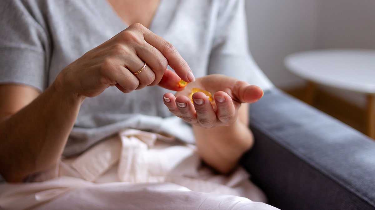 Person taking yellow softgel vitamin or supplement capsules from their hand while sitting on a couch at home.