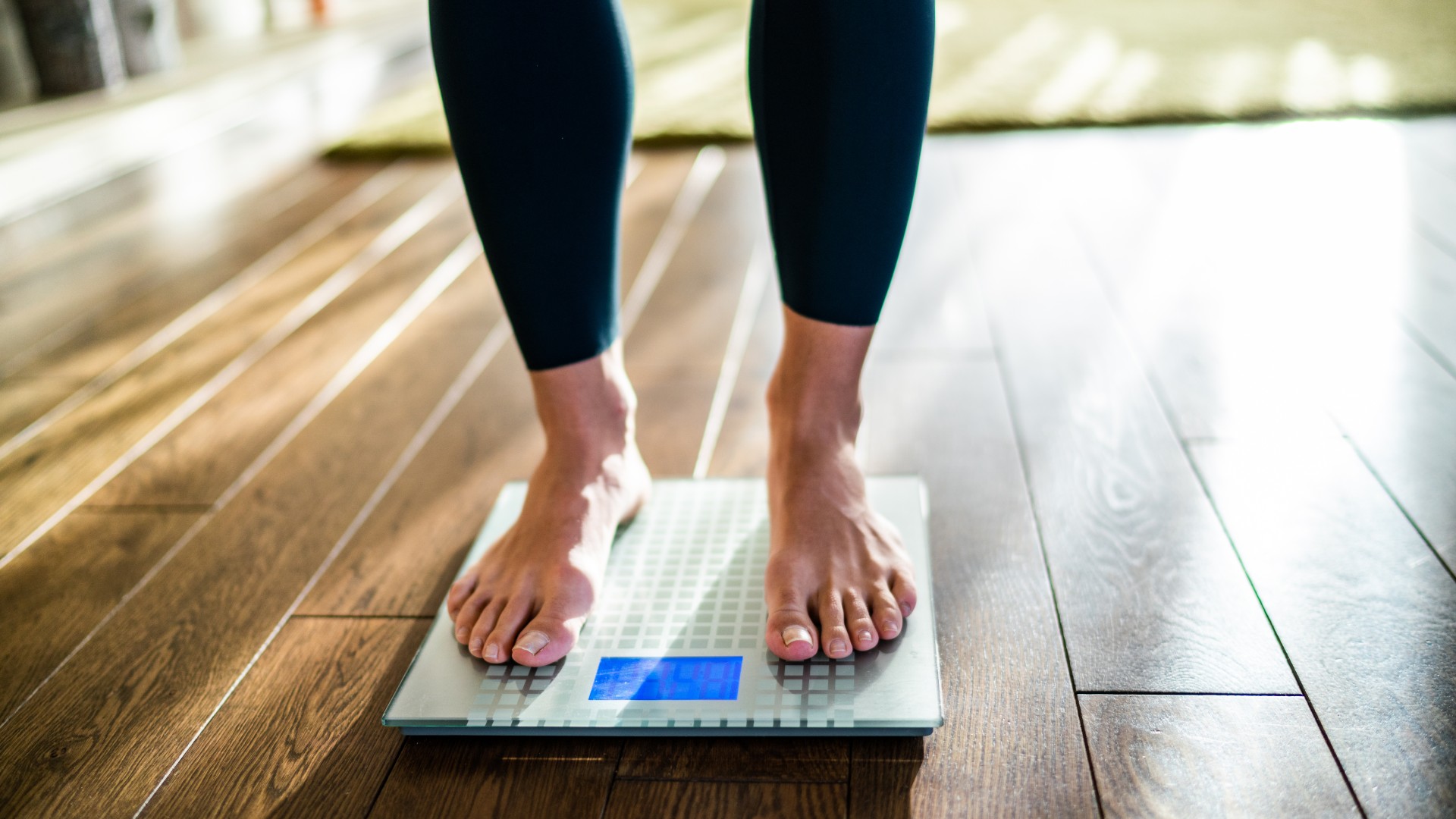 A woman's feet are shown standing on weighing scales that are placed on a wooden floor. She is wearing black leggings and nothing on her feet.