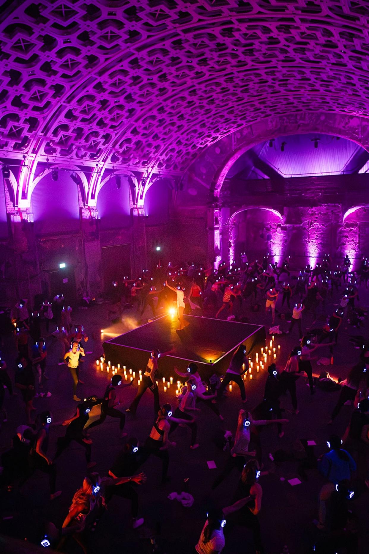 A hall lit only with LED light with a platform in the center and people dancing around it.