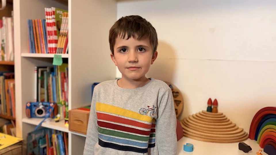 A young child standing indoors in front of shelves of books and wooden toys.