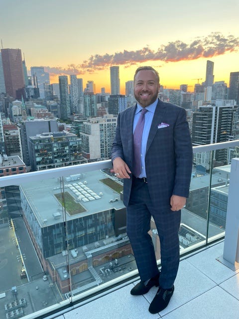 Man in formal attire standing on a rooftop with a city skyline at sunset.