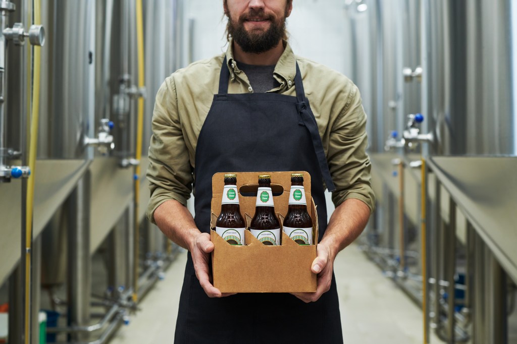 A man in an apron holding a six-pack of "Good Beer" in a brewery.