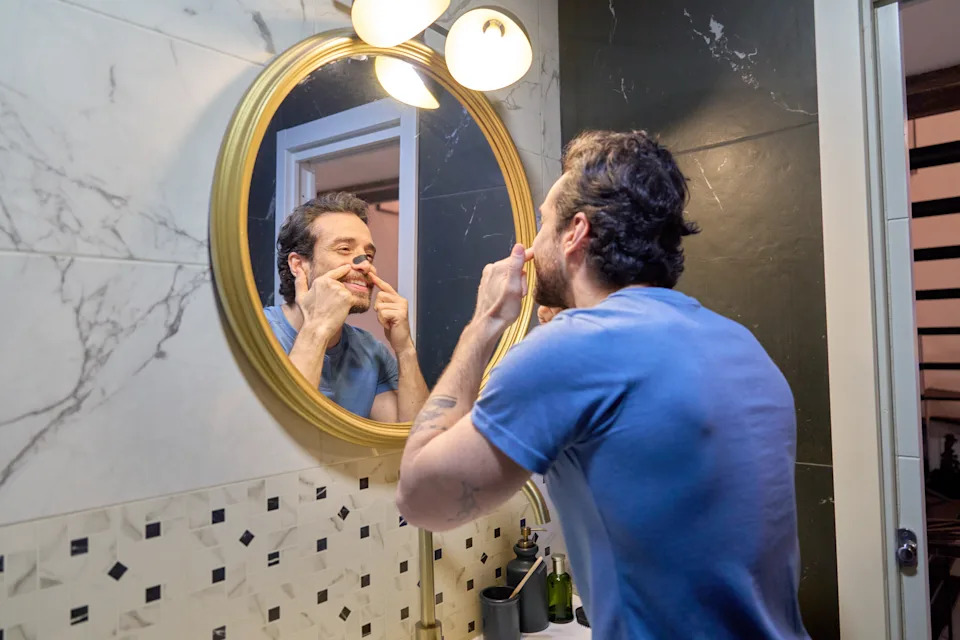 Young adult man smiling, applying a black nose pore strip in front of a mirror, engaging in his morning self-care routine