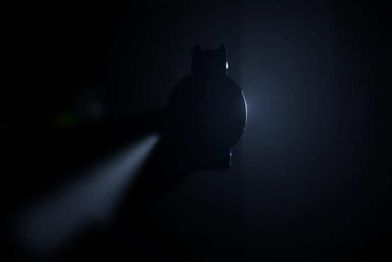 An extreme close-up photograph of a gun trigger mechanism, the metal parts and mechanisms sharply illuminated against a pitch-black background, creating a stark, gritty, investigative aesthetic.