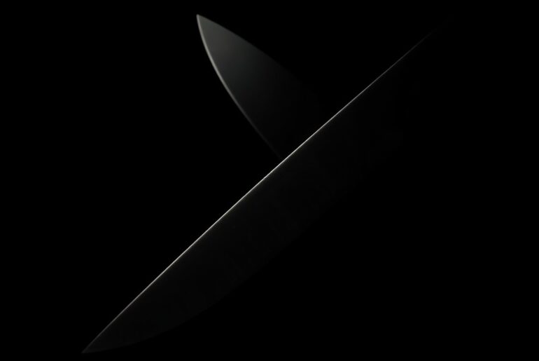 An extreme close-up of a sharp knife blade, the harsh lighting and dark background creating a stark, gritty, and investigative aesthetic to conceptually represent the violent incident at the gym.